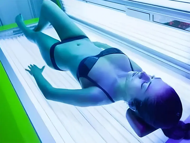 A woman wearing a bikini lies inside an illuminated red light skin care bed with her eyes closed.