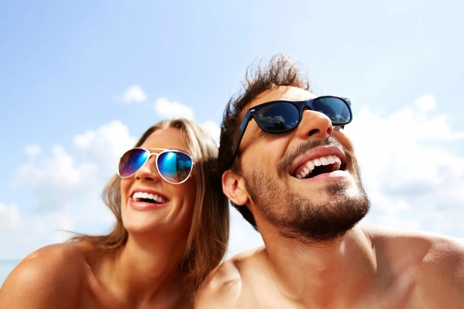 A joyful man and woman wearing sunglasses, smiling under a clear blue sky after their red light skin care session.