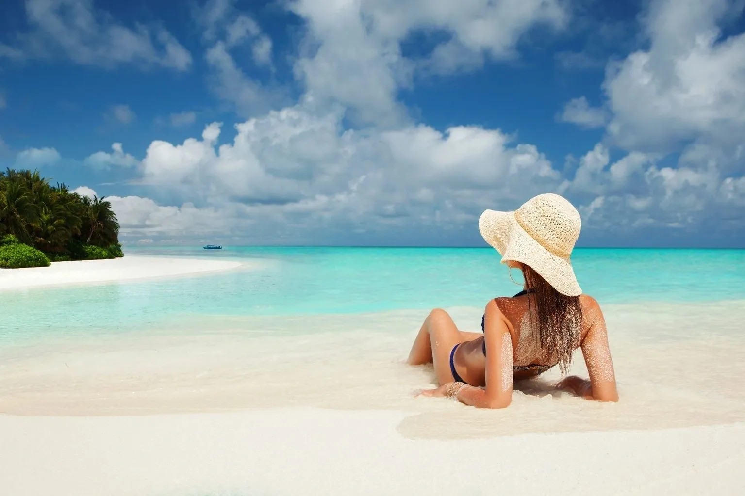 Woman in a straw hat relaxing on a sandy beach, facing a clear blue ocean under a bright sky with her Red Light Skin Care device nearby.